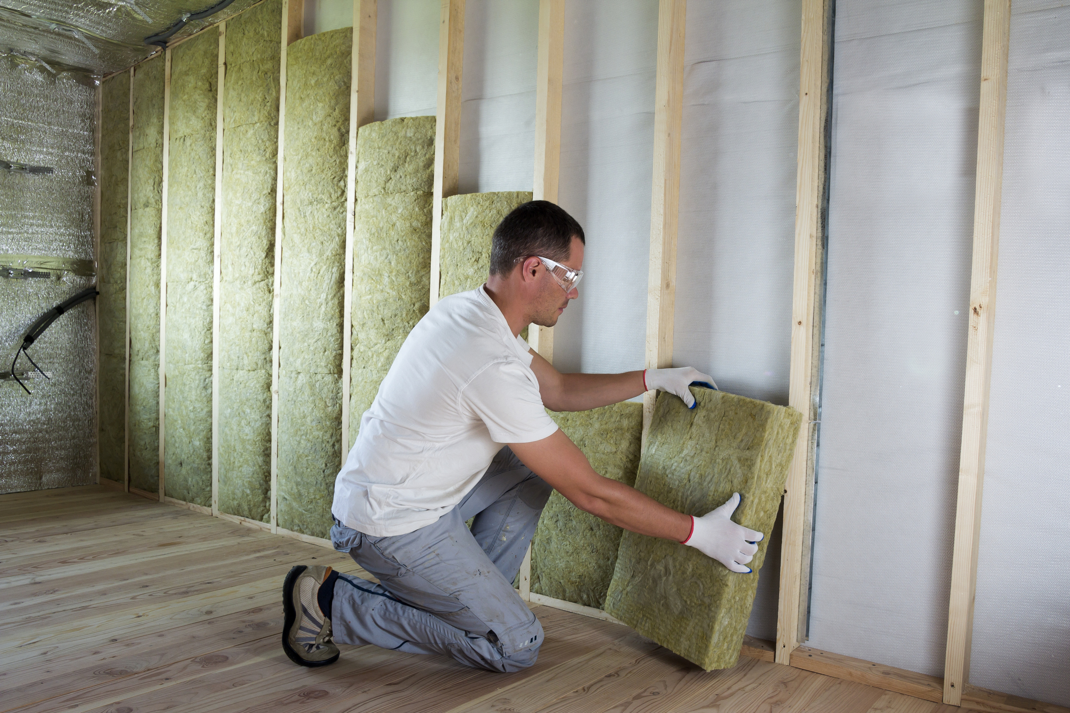Worker in protective goggles and respirator insulating rock wool insulation in wooden frame for future house walls