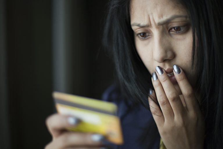 Indoor image of a shocked and worried young woman looking at credit card she is holding and putting her other hand on her face while giving frowning expression on face. One person, horizontal composition with copy space and selective focus.