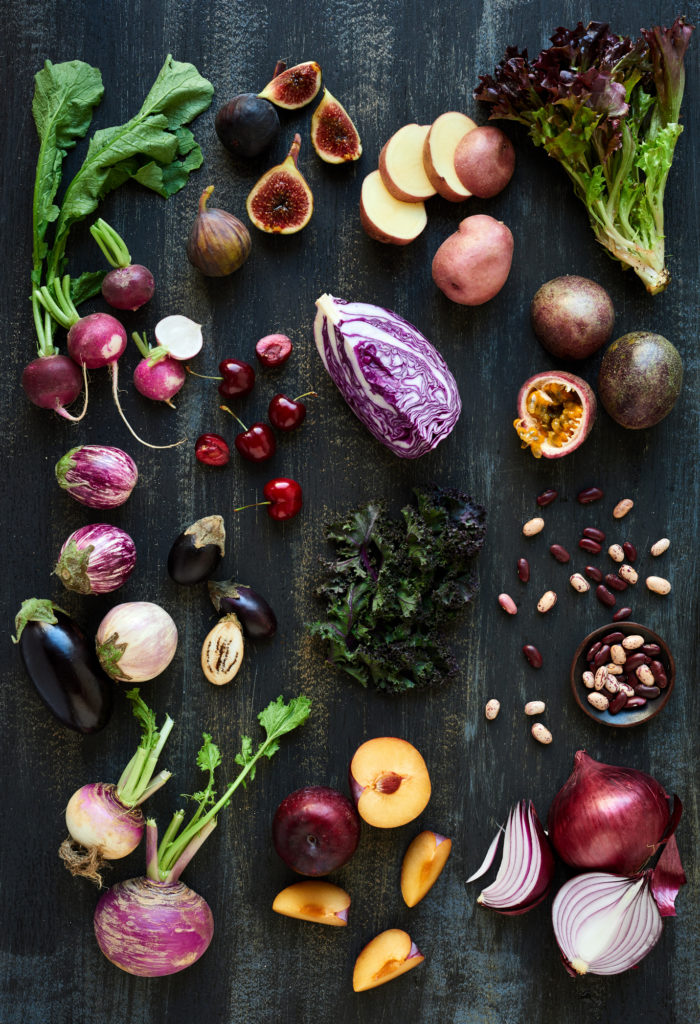 Collection of fresh purple toned vegetables and fruits on dark rustic distressed background, heirloom eggplant, fig, aubergine, cherries, radishes, lettuce, beans passionfruit, cabbage, plum, onion